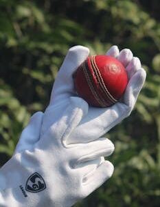 Close-up of a red cricket ball held in white gloves outdoors.